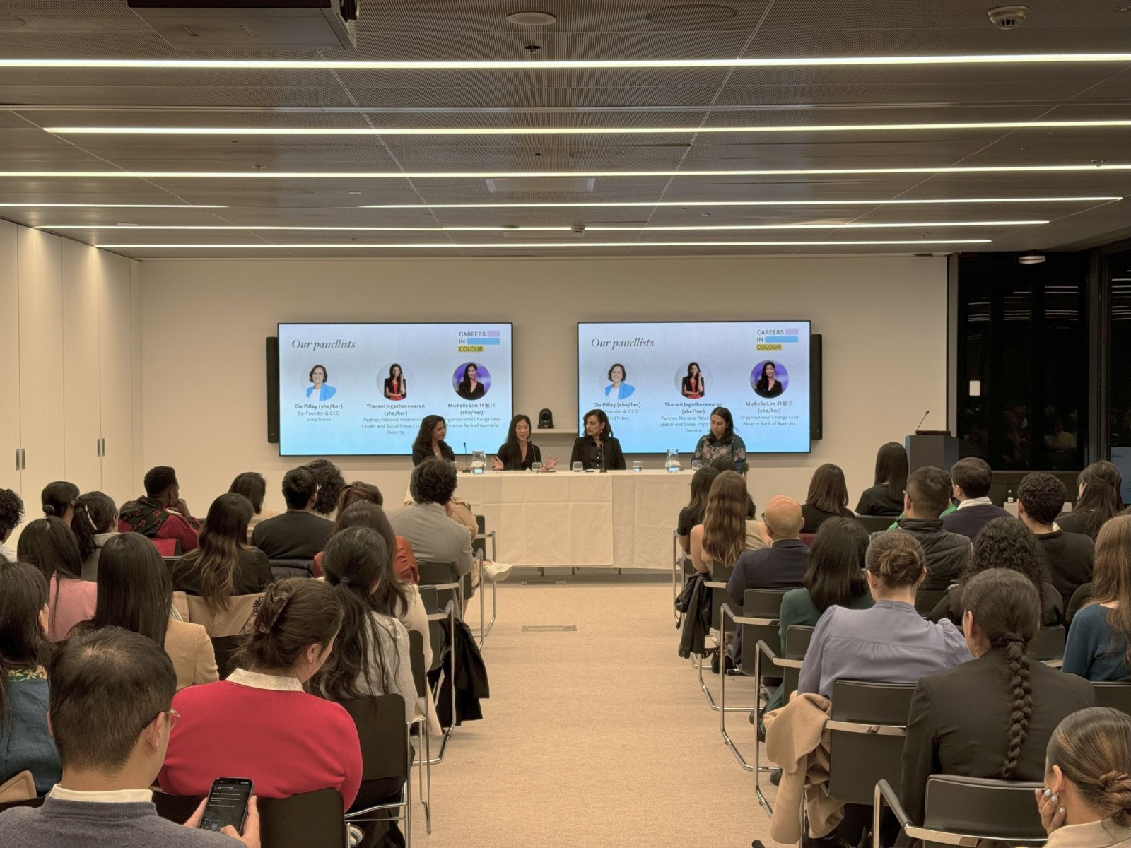 A Careers in Colour panel discussion on workplace racial diversity, featuring four speakers seated at a front table, addressing a diverse audience in a modern conference room with presentation screens displaying panelist details.